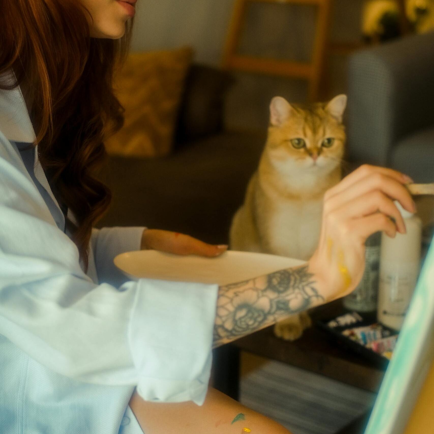 Artist in studio painting alongside her curious feline friend, surrounded by artistic tools.